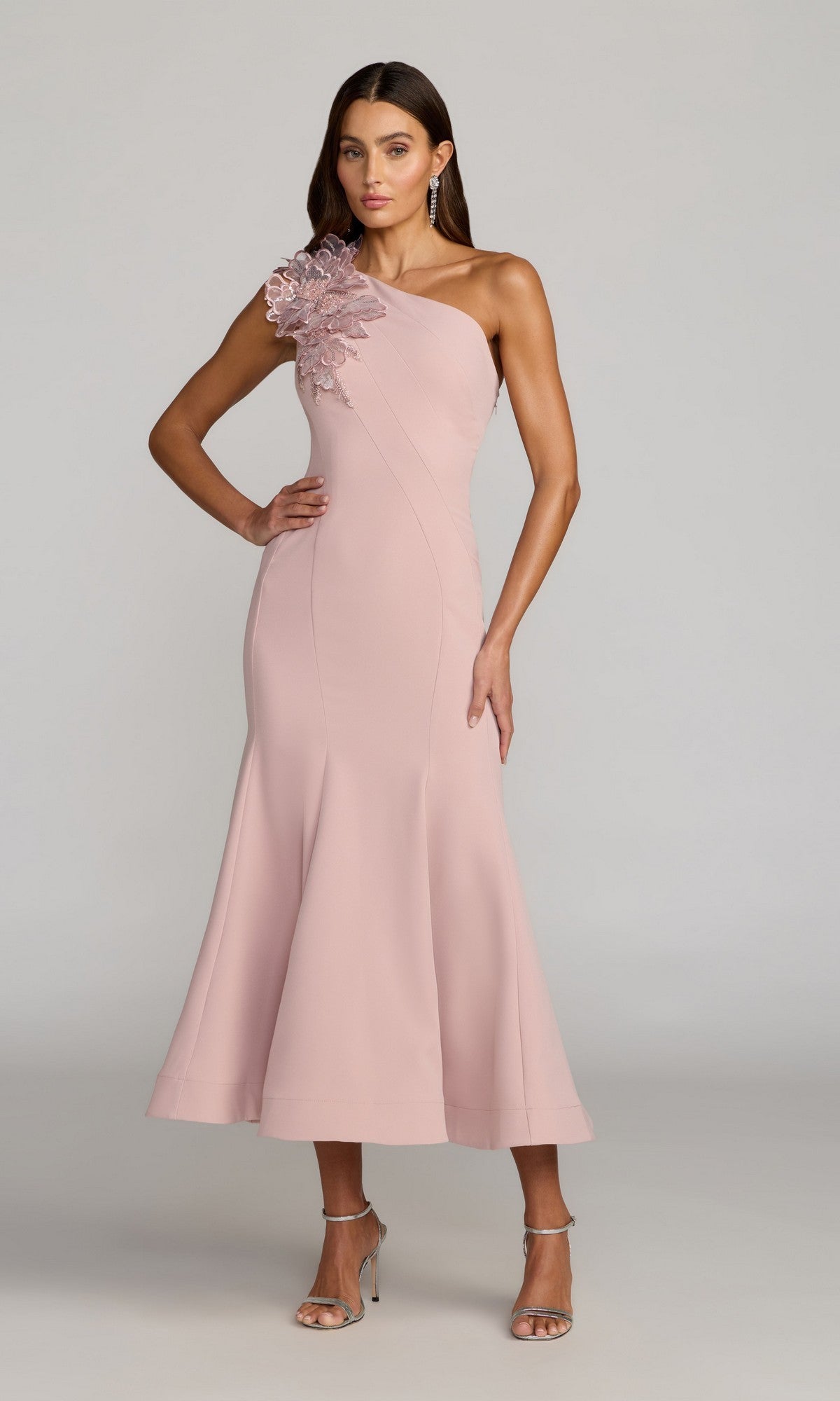A woman stands against a plain light gray background, wearing the Tea-Length Party Dress: Mac Duggal 13222 in rose—a sophisticated one-shoulder crepe midi dress featuring eye-catching 3D floral embellishment on the shoulder.